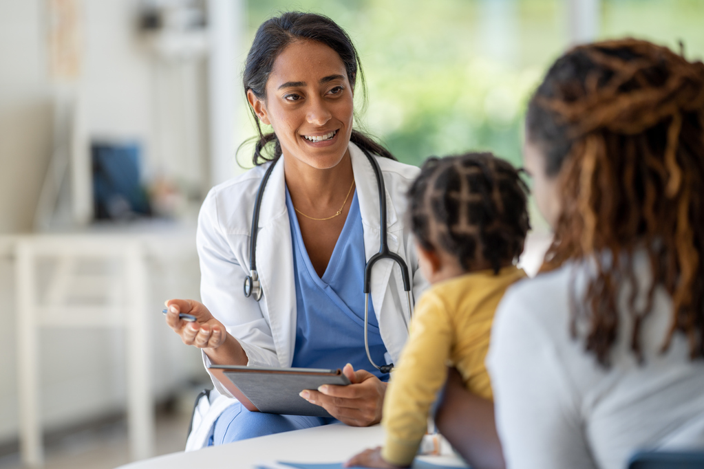 A toddler sits up on an exam table during a routine check-up as his mother holds him securely in place. The female doctor is holding out a tablet as they review some tests together.