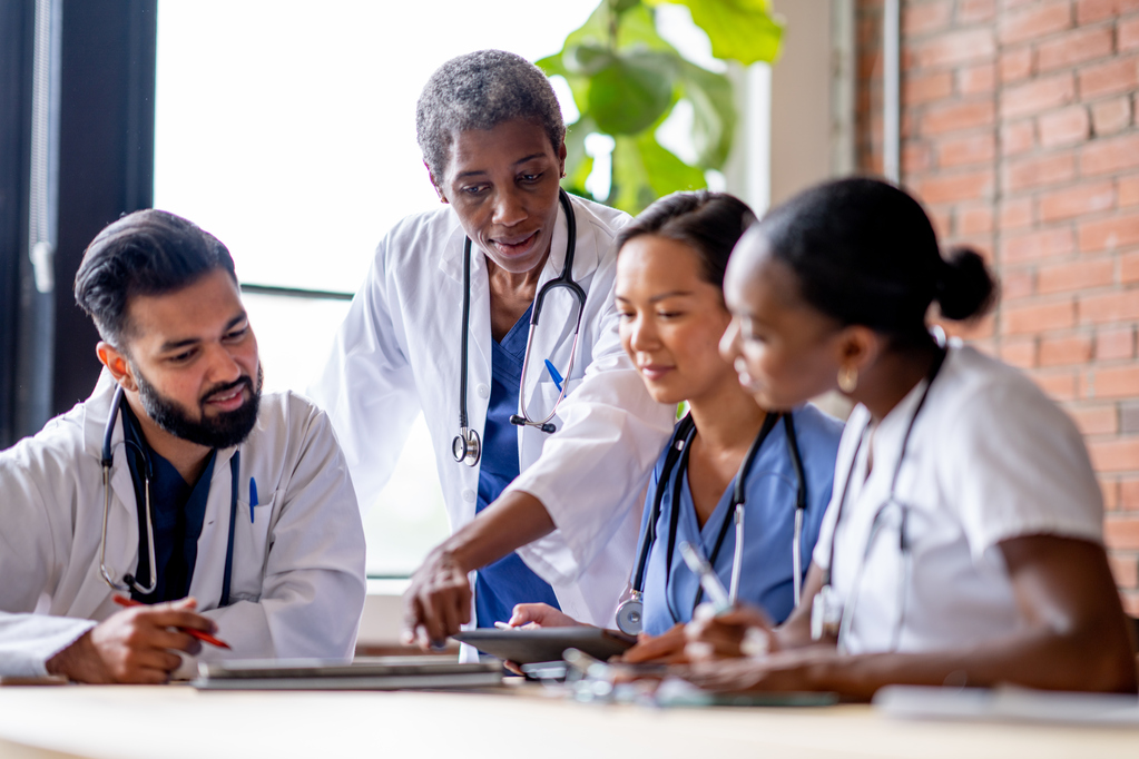 A female doctor stands at the head of a table as she meets with three medical students to review their cases with them. They are each dressed professionally in scrubs and lab coats and are listening attentively as the doctor makes remarks about the files in front of them on the table.