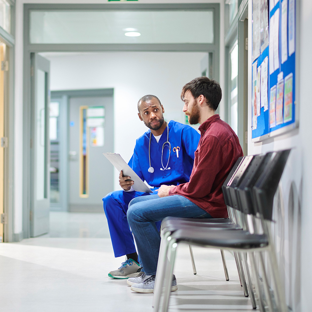 A doctor in scrubs sits with and speaks to a man in a hospital hallway